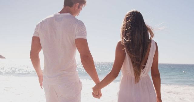 A young Caucasian couple holds hands while walking along a sunny beach, with copy space. Their relaxed posture and the serene coastal setting evoke a sense of romance and tranquility.