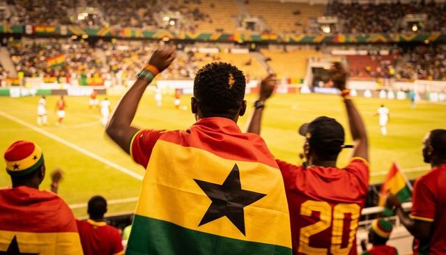 Ghanaian football fans cheering with flag draped over shoulders in night stadium crowd