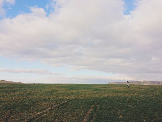 Image showcasing a solitary person strolling across a large green field under a cloudy sky. This serene and open landscape evokes feelings of peace and tranquility. Ideal for use in projects related to relaxation, inspiration, travel, nature, and simplicity. It can be used in blog posts, websites, and promotional materials emphasizing calmness, solitude, and the beauty of wide open spaces.