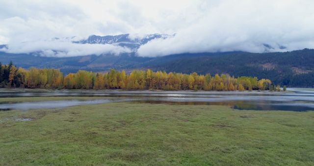 A tranquil autumn scene featuring a calm lake surrounded by trees with colorful foliage and a mountain range in the background partially covered by clouds. Great for travel blogs, nature conservation articles, fall-themed promotions, and environmental awareness campaigns.