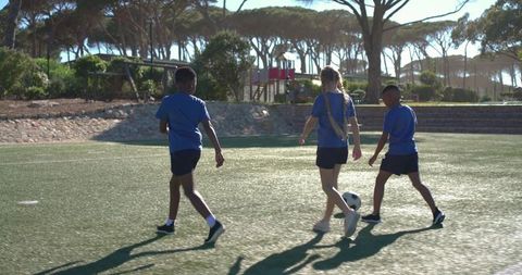 Multiracial teens walking on soccer field dribbling ball in sunlight showing teamwork