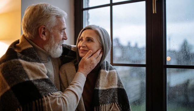 Senior couple sharing cozy plaid blanket by rainy window, embracing warmth and intimacy