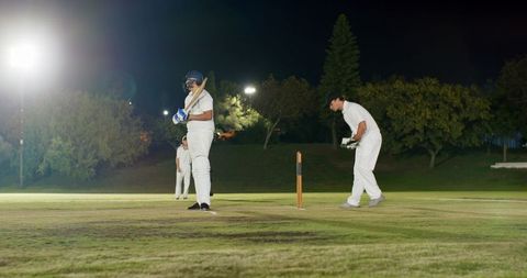 Night cricket match under floodlights with batsman, wicketkeeper and fielder on grass