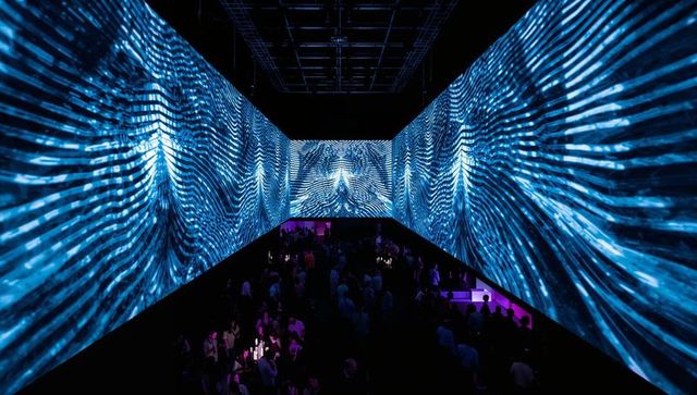 Immersive three-wall projection installation with kinetic blue wave visuals and crowd silhouettes