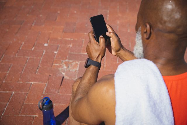 Senior man sitting on brick pavement using smartphone after exercise. He has a towel over his shoulder and a water bottle nearby, indicating a focus on fitness and hydration. This image is ideal for promoting active senior lifestyles, fitness apps, health and wellness products, or technology use among older adults.