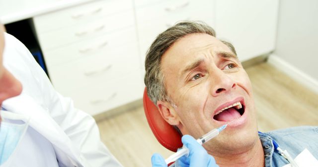 Adult man looking anxious while receiving dental injection from dentist with gloves. Use for topics related to dental care, medical procedures, patient anxiety, and health insurance.