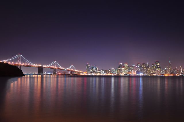San Francisco Bay Bridge extending over water towards illuminated city skyline at night. Reflective water creates scenic view of urban architecture under clear night sky. Ideal for travel brochures, city skyline photos, urban themes, California tourism materials or website headers.