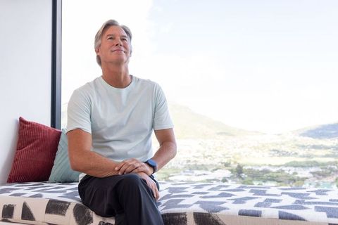 Mature man sitting by large window gazing at sunlit valley, serene modern interior lifestyle