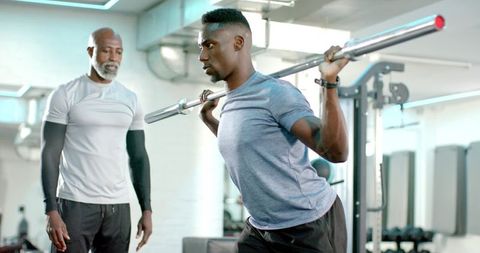 African american personal trainer coaching young man performing barbell squat in modern gym