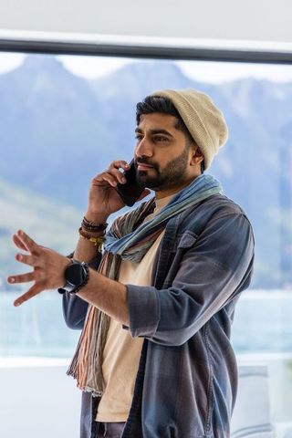 Indian traveler talking on phone at panoramic window with lake and mountain view