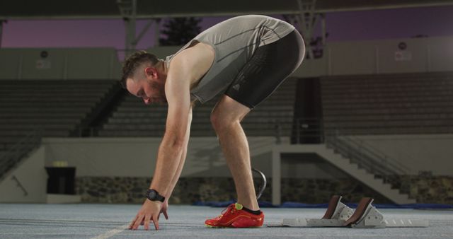 Male athlete performing pre-run stretching exercises on a track in a stadium. He is wearing a grey tank top, black shorts, and red athletic shoes. The image captures the intimacy of preparation before a sprint or run, making it ideal for use in fitness campaigns, sports advertisements, or motivational materials for athletes and runners.