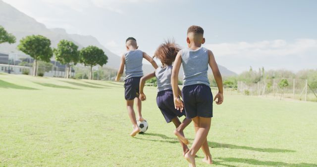 Three children are playing soccer barefoot on a sunny day in an open field. They are wearing similar casual sportswear, indicating team spirit or shared activity. This image is perfect for promoting outdoor activities, children's sports programs, summer camps, or teamwork and friendship themes.