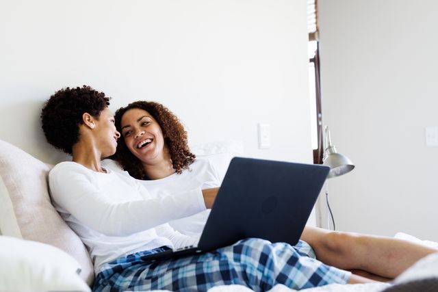 This image shows a happy, diverse lesbian couple sitting on a bed, looking at a laptop together and laughing. Ideal for use in articles or advertisements about relationships, LGBTQ+ representation, home life, technology use in daily life, and promoting inclusivity and diversity.