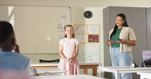 Young girl presenting to class with supportive teacher holding tablet in modern classroom