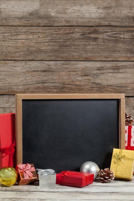 Christmas decorations and various gift boxes are arranged around a blank chalkboard on a wooden surface. Pine cones and festive ornaments add to the holiday atmosphere. This setup can be used for Christmas party invitations, holiday announcements, seasonal promotions, or festive greeting cards.