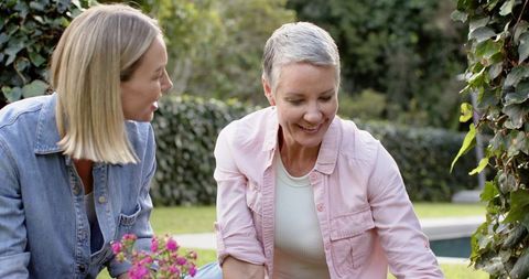 Two women gardening by pool tending pink potted flowers, smiling and kneeling in backyard
