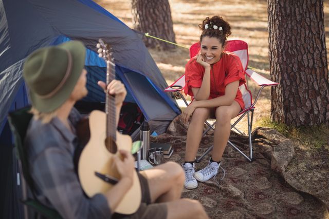 Happy young looking at her boyfriend playing guitar by tent at forest