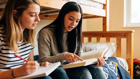Two teen girls studying together in dorm room pointing to textbook and taking notes