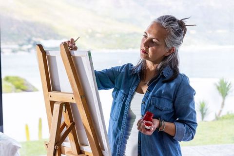 Senior woman painting at easel in bright coastal studio holding brush and red paint