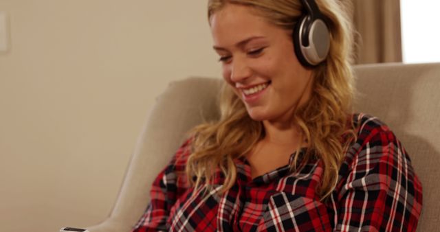 Young woman wearing headphones and smiling while using mobile device. Ideal for themes of relaxation, leisure, technology use and casual lifestyle. Suitable for illustrating concepts of enjoying music or engaging in technology.