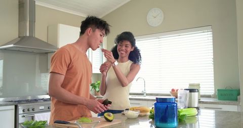 African american woman feeding avocado to asian man while preparing breakfast on kitchen island