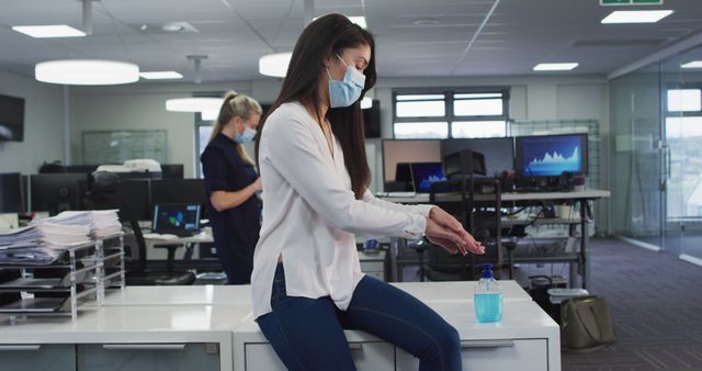 Workers in an office environment practicing COVID-19 safety measures by wearing masks and sanitizing their hands. Ideal for illustrating health and safety protocols in the workplace, pandemic precautions, and corporate responsibility toward employee health.