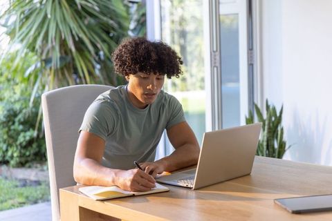 African american man working remotely at bright home desk writing notes and using laptop