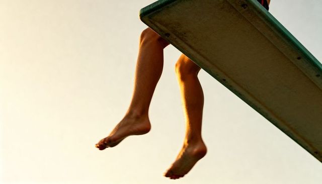 Dangling legs bare feet sitting on green diving board at golden hour