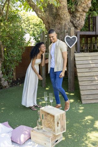 Couple celebrating backyard proposal with marry me heart sign, rustic crates and champagne