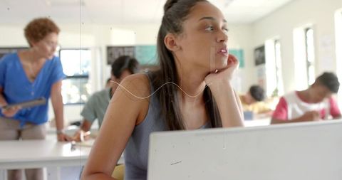 Pensive teenage student resting chin on hand studying with laptop in sunlit classroom