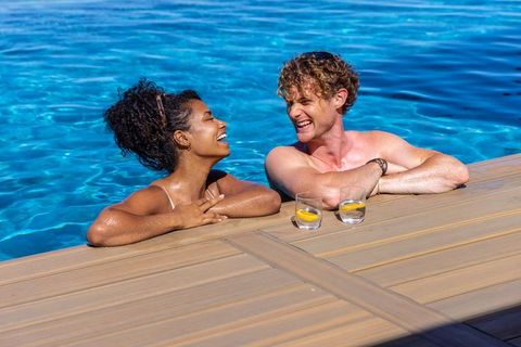 Diverse couple relaxing at pool edge, smiling and chatting over lemon drinks on deck