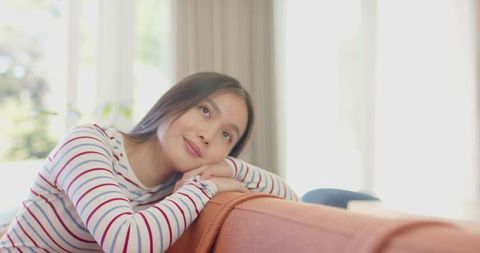Asian woman dreamily resting on rust-colored sofa in sunlit living room