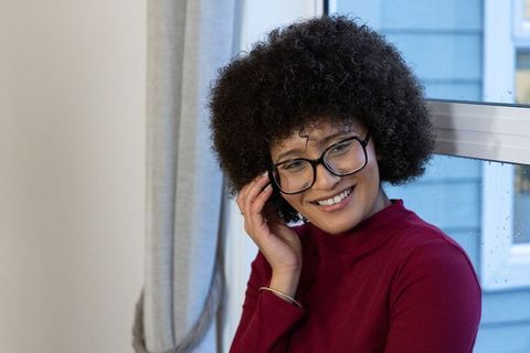 African american woman smiling while adjusting glasses by window on rainy day in burgundy turtleneck