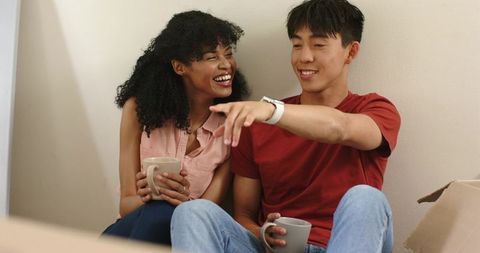 Young diverse couple laughing and pointing while unpacking in cozy new home with mugs