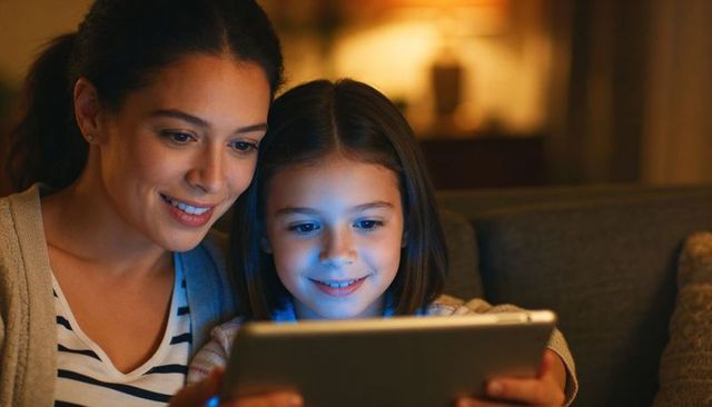 Mother and daughter sharing tablet on couch, evening family bonding warm glow digital learning