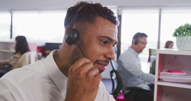 Young man with headset in modern office environment providing customer support while engaging with clients over the phone. Colleagues working in the background, suggesting teamwork and a professional business setting. Ideal for use in content related to customer service, office work, business communication, teamwork, and professional environments.