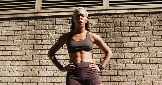 This image shows a fit woman standing confidently against a brick wall in minimal sportswear, looking determined and ready for an intense workout. Ideal for use in fitness, health and wellness blogs, advertising athletic wear, motivational posters, or content focused on women's empowerment and strength.