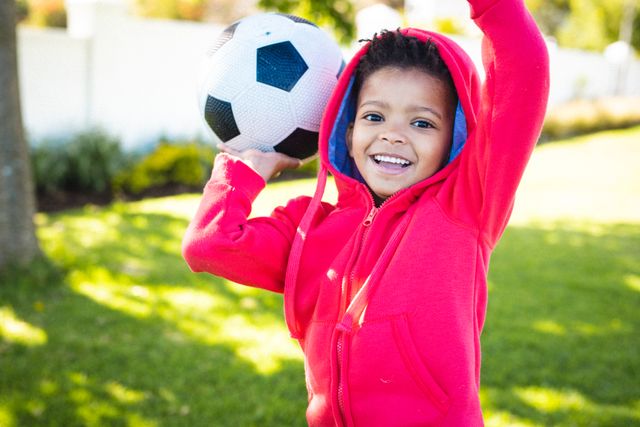 Young African American boy smiling and holding a soccer ball in a backyard. Ideal for use in advertisements, blogs, and articles about childhood, outdoor activities, family time, and sports. Perfect for promoting children's clothing, sports equipment, and family-oriented products.