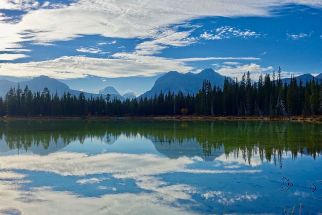 Reflection of mountains and tall pine trees in clear, calm lake under partly cloudy sky. Ideal for travel blogs, nature posters, calendar backgrounds, meditation aids, and outdoor adventure promotions.