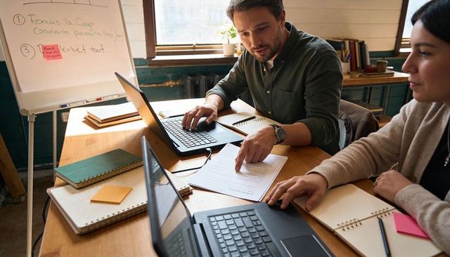 Man pointing at document while collaborating with colleague over laptops in modern workspace