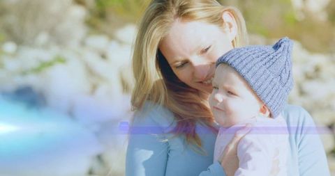 Sunlit mother cradling baby girl in blue beanie outdoors with gentle lens flare