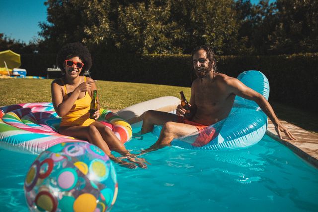 Diverse couple enjoying a sunny day in the pool, sitting on inflatable chairs, and drinking beer. Ideal for use in summer vacation promotions, leisure and lifestyle blogs, or advertisements for outdoor activities and poolside products.