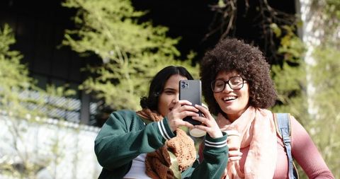 College friends taking selfie on sunny campus, scarves and backpacks, holding coffee and smiling