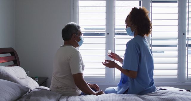 Nurse giving medication to elderly patient in a bright room with natural light filtering through windows. Both wear face masks, indicating a concern for health and safety, possibly due to a pandemic or other contagious conditions. Can be used for articles, blogs, or educational materials focusing on elderly care, home healthcare services, or health and safety practices.