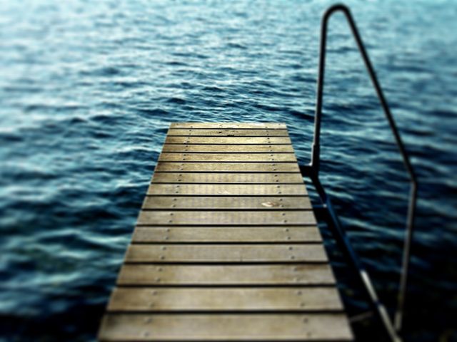 Wooden pier extending into serene, calm blue water. Ideal for themes of relaxation, nature, summer vacations, outdoor activities, and peaceful landscapes. Could be used for advertisements promoting travel, getaways, or wellness.