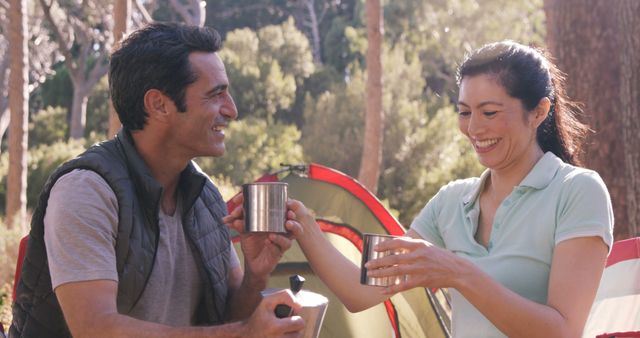 Image shows a happy couple enjoying coffee in a forest setting, with a tent in the background. Smiling and engaging in leisure outdoor activity. Useful for depicting camping trips, nature getaways, and outdoor relaxation.