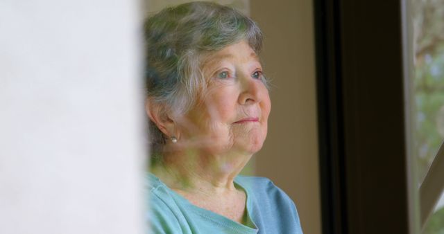 Senior Caucasian woman looks thoughtfully out a window, with copy space. Her expression suggests reflection or anticipation at home.