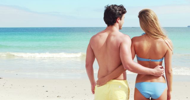 Couple standing on sandy beach, embracing while looking at the ocean. Ideal for travel promotion, vacation packages, romantic getaways, beachwear advertisements, and relaxation-themed content.