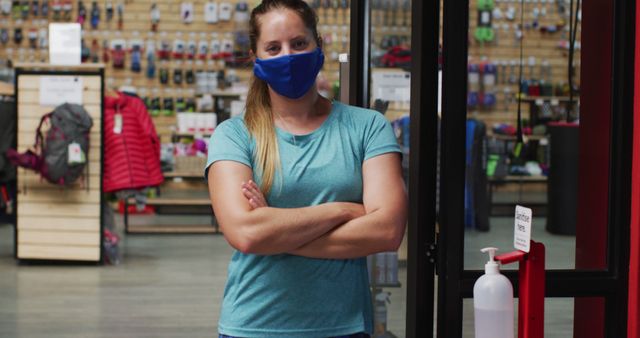 Caucasian female shopkeeper wearing face mask in the doorway of sports shop with arms crossed. fitness and leisure retail during coronavirus covid 19 pandemic.