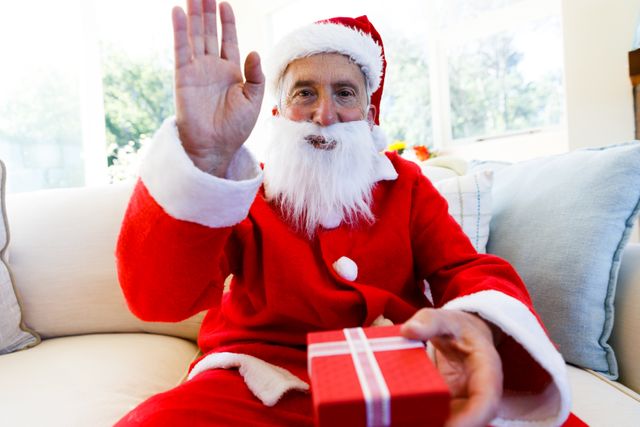 Front view of an old caucasian man wearing a santa clause costume holding out a christmas gift in front of him with his other hand waving. he is sitting on the couch in the living room.